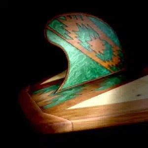 Close-up studio photography of the fin for a wooden paddleboard by Little Bay Boards, showcasing rich natural wood textures and a deep forest green chevron art inlay over a solid black backdrop.