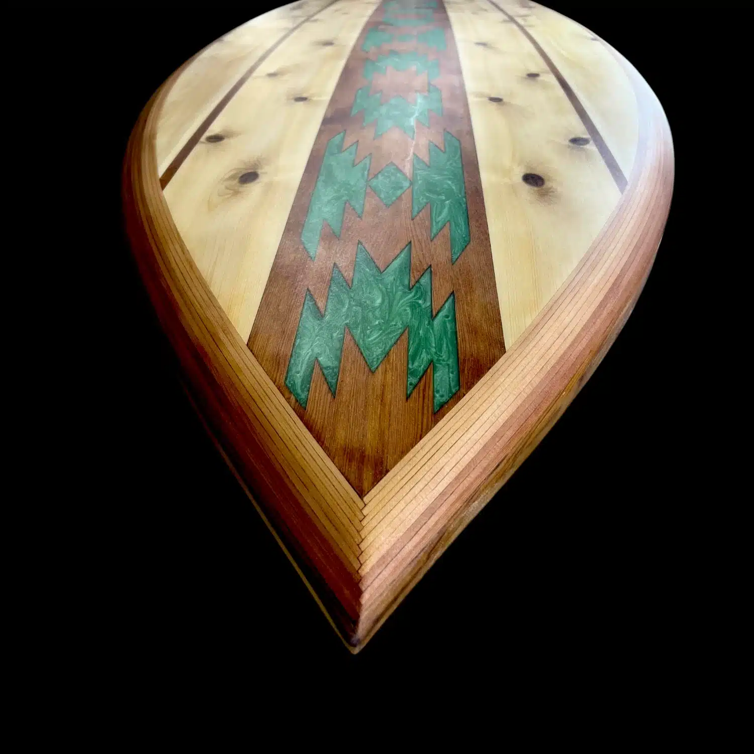 Close-up studio photography of the nose on a custom wooden paddleboard by Little Bay Boards, showcasing rich natural wood textures and a deep forest green chevron art inlay over a solid black backdrop.